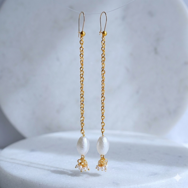 Pair of dainty long gold earrings with white pearls hanging over a marble pedestal.